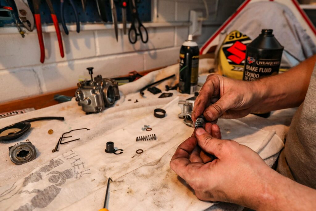 Close-up of hands repairing a carburetor with tools on a workbench in a garage setting.